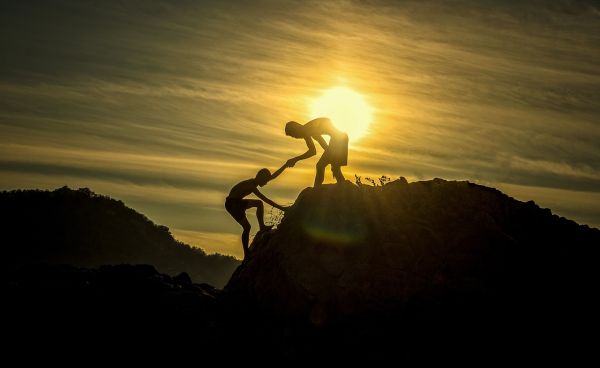one person helping another reach the top of a mountain at sunset